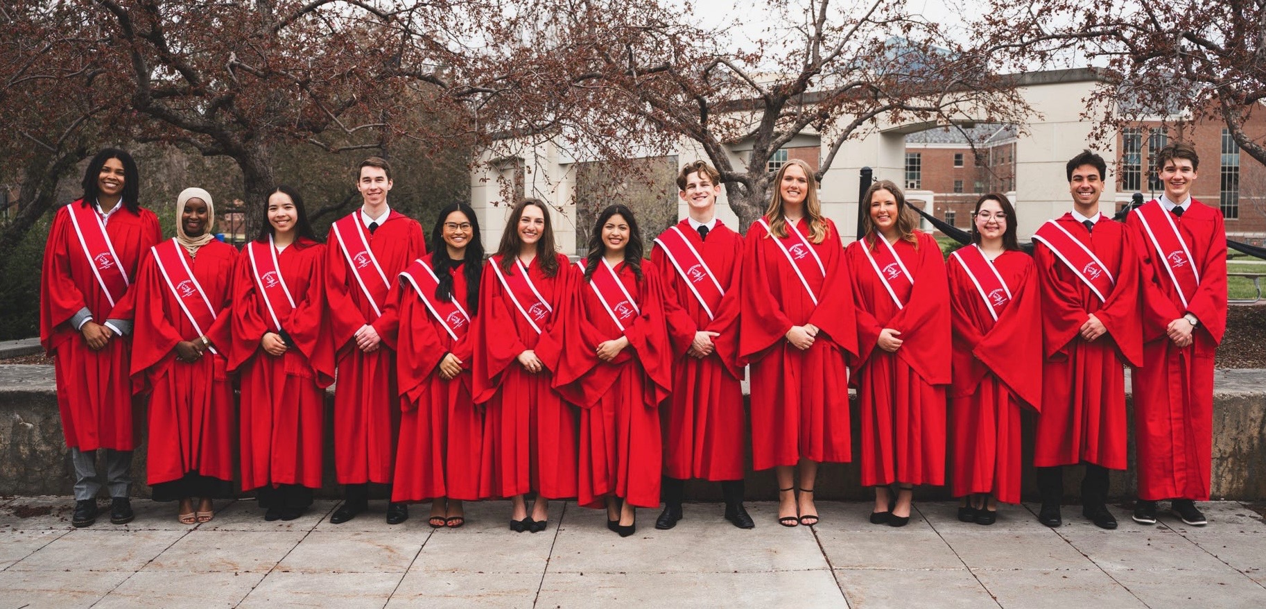A group of students in red graduation robes standing in a row outdoors, smiling.